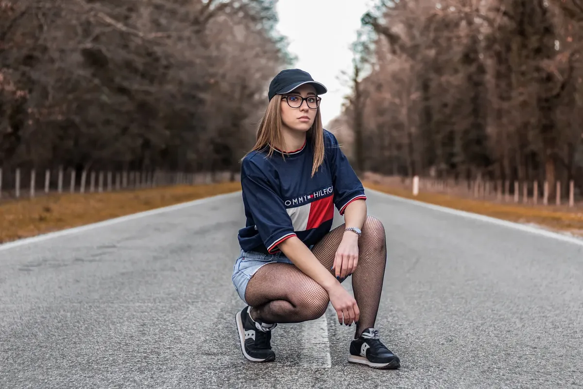 Stylish woman in casual attire kneeling on an empty forest road, showcasing modern fashion.